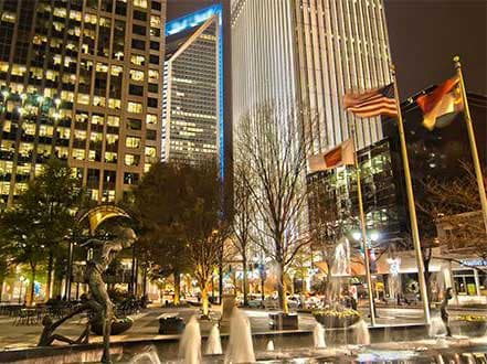 Vibrant city skyline at night with tall buildings, flags, and a fountain in the foreground.